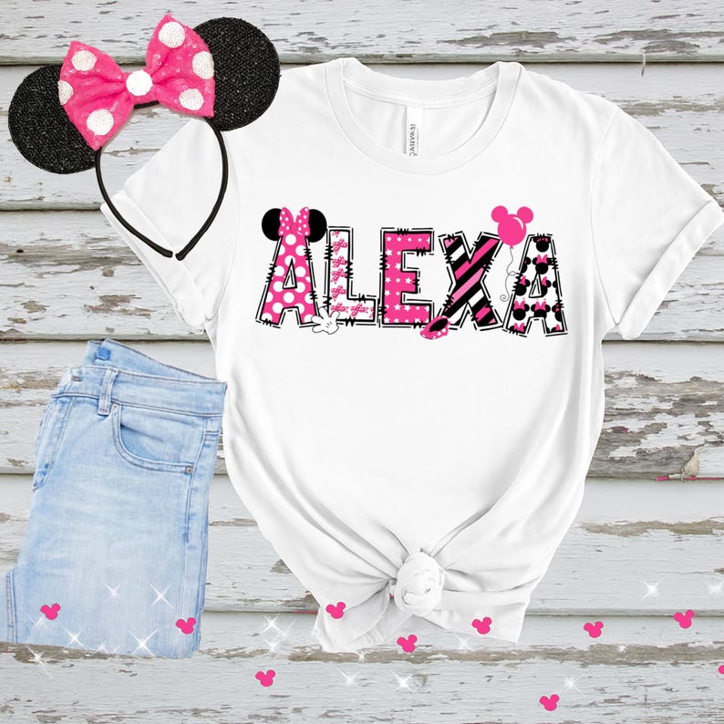 K&ouml;nnte beinhalten: Wei&szlig;es T-Shirt mit dem Namen "ALEXA" in rosa und schwarz gemusterten Buchstaben, mit Minnie-Mouse-Akzenten. Ein passendes rosa gepunktetes Schleifen-Stirnband und hellblaue Jeans sind ebenfalls im Bild.