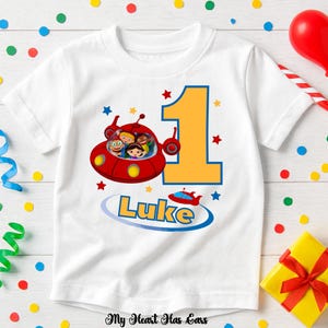 May include: White short-sleeved t-shirt featuring a red spaceship graphic with cartoon characters, the number 1 in yellow, and the name "Luke" in blue. The design includes stars and a small red spaceship.