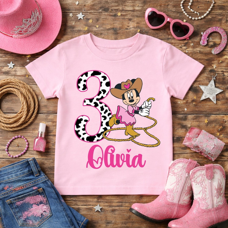 May include: A pink t-shirt featuring a cartoon Minnie Mouse dressed as a cowgirl, holding a rope. The number "3" is in a black and white cow print, with the name "Olivia" below. Accessories include pink boots, a hat, and sunglasses.