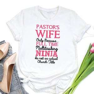 May include: White t-shirt with the text "PASTOR'S WIFE Only Because... FULL TIME Multitasking NINJA Is not an actual Church Title" in pink and black lettering. The shirt is folded, with a pair of shoes and flowers in the background.