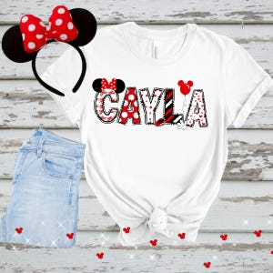 以下が含まれることがあります： 赤、黒、白のデザインで「CAYLA」という名前が入った白いTシャツ、ミニーマウスの耳のヘッドバンド、ジーンズ。名前には水玉模様、ハート、ストライプが使われています。赤いミッキーマウスの頭の形がジーンズの周りに散らばっています。