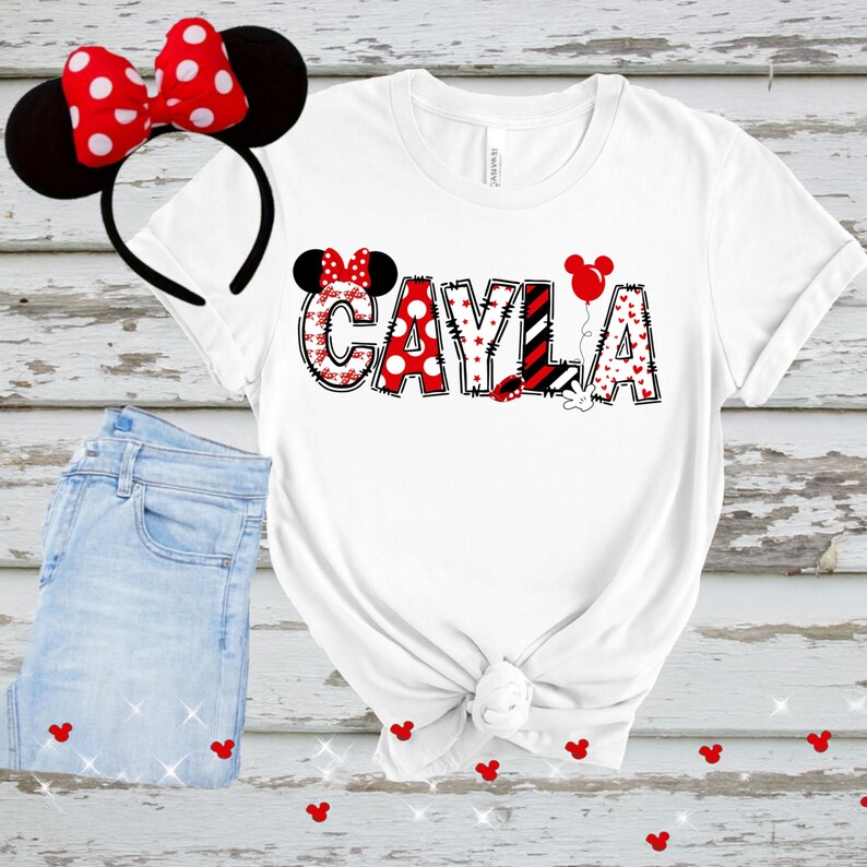以下が含まれることがあります： 赤、黒、白のデザインで「CAYLA」という名前が入った白いTシャツ、ミニーマウスの耳のヘッドバンド、ジーンズ。名前には水玉模様、ハート、ストライプが使われています。赤いミッキーマウスの頭の形がジーンズの周りに散らばっています。