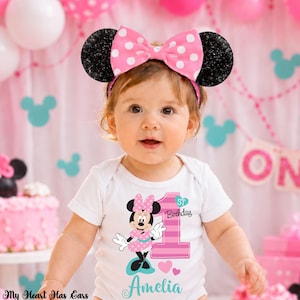 K&ouml;nnte beinhalten: Ein Baby tr&auml;gt einen wei&szlig;en Body mit einem Minnie-Maus-Motiv und dem Text "1. Geburtstag" und "Amelia". Das Baby tr&auml;gt auch ein rosa Schleifen-Stirnband mit schwarzen Glitzerohren. Rosa und wei&szlig;e Luftballons sind im Hintergrund.