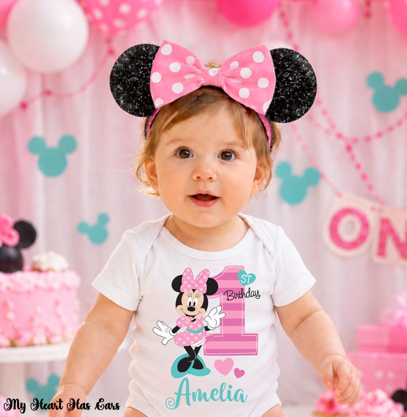 K&ouml;nnte beinhalten: Ein Baby tr&auml;gt einen wei&szlig;en Body mit einem Minnie-Maus-Motiv und dem Text "1. Geburtstag" und "Amelia". Das Baby tr&auml;gt auch ein rosa Schleifen-Stirnband mit schwarzen Glitzerohren. Rosa und wei&szlig;e Luftballons sind im Hintergrund.