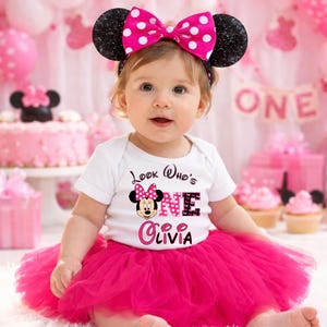 May include: A baby wearing a white onesie with a Minnie Mouse graphic and the text "Look Who's ONE Olivia". The baby is wearing a pink tutu and a Minnie Mouse ear headband with a pink bow. Pink balloons and a cake are in the background.