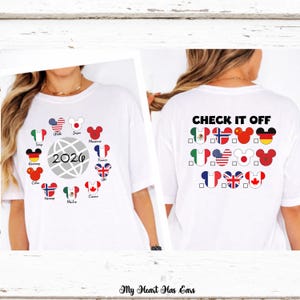 Könnte beinhalten: Ein weißes T-Shirt mit einem Grafikdesign, das einen Globus und das Jahr 2026 zeigt, umgeben von Mickey-Maus-förmigen Symbolen mit verschiedenen Nationalflaggen. Die Rückseite zeigt den Text CHECK IT OFF mit einer Checkliste aus Symbolen.