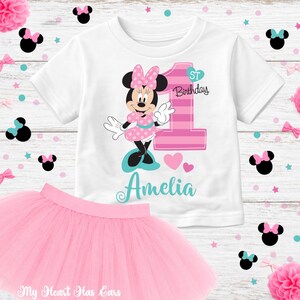 K&ouml;nnte beinhalten: Ein wei&szlig;es T-Shirt mit Minnie Mouse-Design, ein rosa Tutu-Rock und dekorative Elemente. Das Shirt zeigt Minnie Mouse, die Zahl 1 und den Namen "Amelia". Der Tutu-Rock ist hellrosa und perfekt f&uuml;r eine erste Geburtstagsfeier.