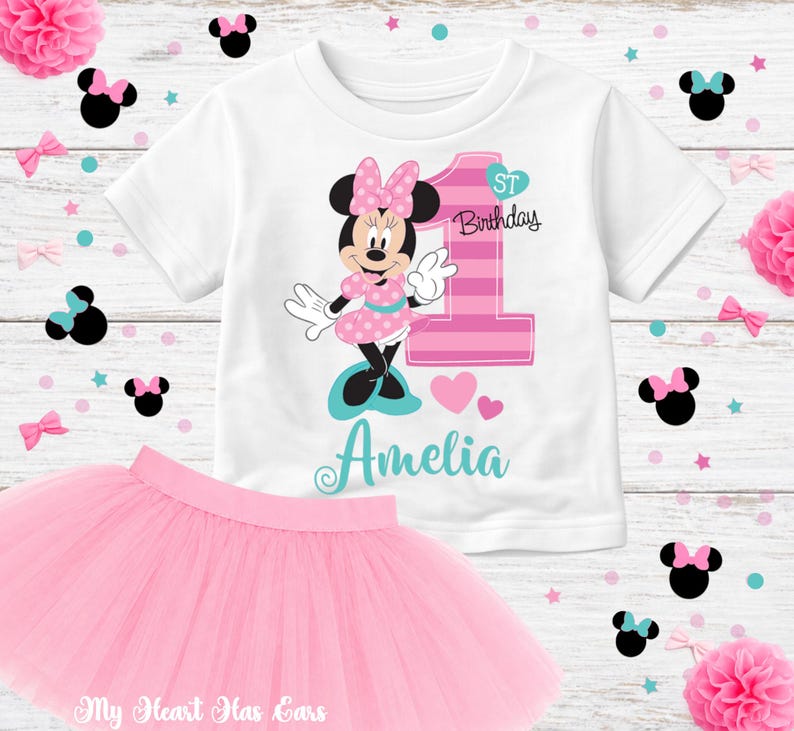 K&ouml;nnte beinhalten: Ein wei&szlig;es T-Shirt mit Minnie Mouse-Design, ein rosa Tutu-Rock und dekorative Elemente. Das Shirt zeigt Minnie Mouse, die Zahl 1 und den Namen "Amelia". Der Tutu-Rock ist hellrosa und perfekt f&uuml;r eine erste Geburtstagsfeier.