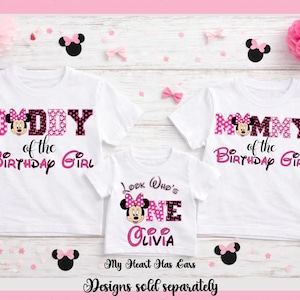May include: Three white t-shirts with Minnie Mouse designs. One says "Daddy of the Birthday Girl", another says "Mommy of the Birthday Girl", and the third says "Look Who's One Olivia". Pink and black accents.