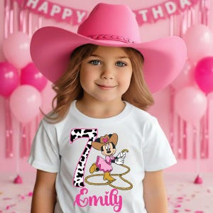 May include: A young girl wearing a pink cowboy hat and a white t-shirt with a Minnie Mouse cowgirl design. The shirt features the number 7 in a cow print pattern and the name "Emily" in pink script. Pink balloons and a "Happy Birthday" banner are in the background.