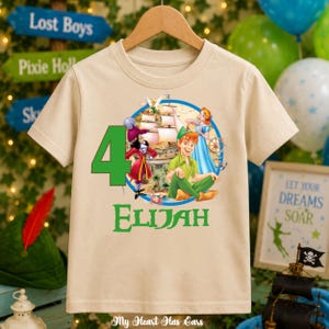 Puede incluir: Una camiseta beige de manga corta con un gráfico de personajes de Peter Pan, un barco pirata, un número 4 grande en verde y el nombre Elijah en texto verde. El diseño está centrado en la parte delantera de la camiseta sobre una percha.