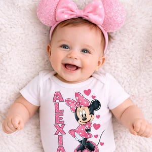 May include: A smiling baby wearing a pink Minnie Mouse ear headband with a matching bow. The baby is wearing a white onesie with the name "ALEXIA" and a Minnie Mouse graphic. Pink hearts are scattered around the Minnie Mouse graphic.