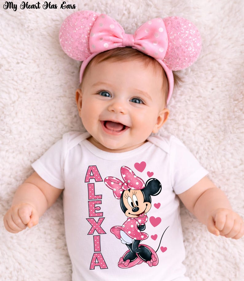 May include: A smiling baby wearing a pink Minnie Mouse ear headband with a matching bow. The baby is wearing a white onesie with the name "ALEXIA" and a Minnie Mouse graphic. Pink hearts are scattered around the Minnie Mouse graphic.