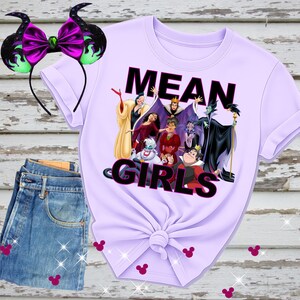 K&ouml;nnte beinhalten: Ein helllila T-Shirt mit dem Text "MEAN GIRLS" und einer Grafik von Disney-B&ouml;sewichten. Ein passendes Stirnband mit lila Schleife und schwarzen H&ouml;rnern befindet sich links. Blaue Jeans sind darunter gefaltet.