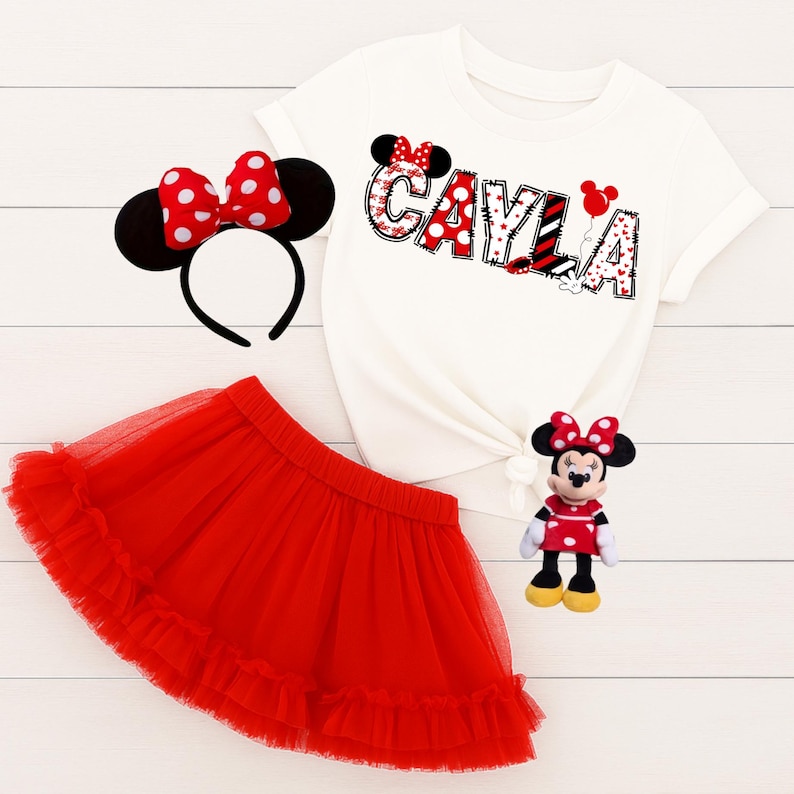 以下が含まれることがあります： 赤と黒のドット柄で「CAYLA」という名前が入った白いTシャツ、赤いチュールスカート、ミニーマウスのヘッドバンド、ミニーマウスのぬいぐるみが入った子供用衣装です。