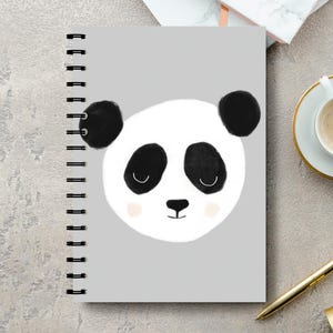 May include: A grey spiral-bound notebook with a panda face design. The panda has black ears, eyes, and nose, with a white face and pink cheeks. The notebook's spiral binding is visible on the left side. The design is simple and cartoon-like.