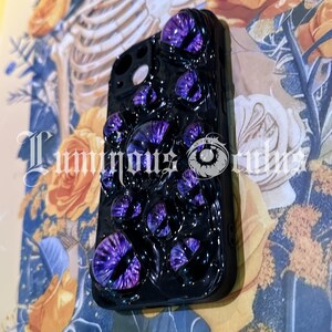 May include: Black phone case adorned with multiple purple eye-shaped embellishments. The case has a glossy finish and features the text "Luminous Orbs". The background includes floral and skeletal imagery.