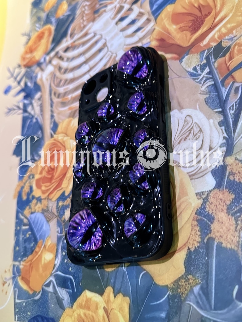 May include: Black phone case adorned with multiple purple eye-shaped embellishments. The case has a glossy finish and features the text "Luminous Orbs". The background includes floral and skeletal imagery.