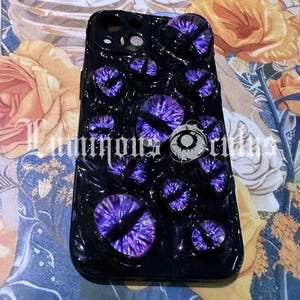 May include: Black phone case featuring multiple purple and black eye designs. The case has a glossy finish and the text "Luminous Brides" is visible. The case is on a floral background.