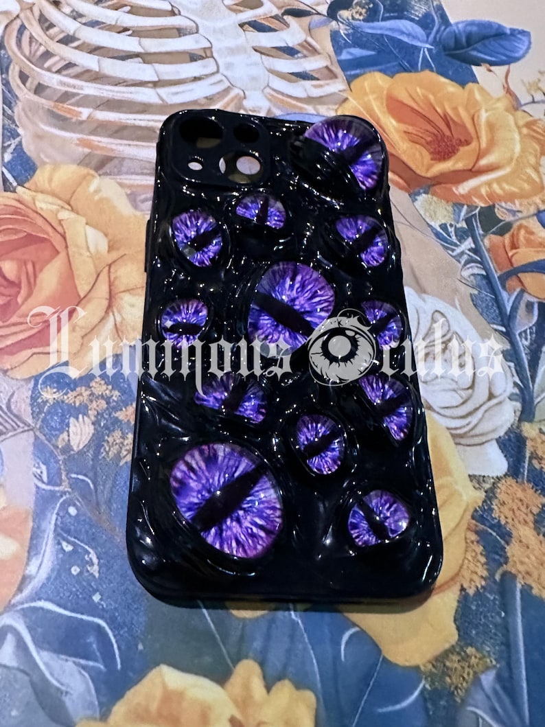 May include: Black phone case featuring multiple purple and black eye designs. The case has a glossy finish and the text "Luminous Brides" is visible. The case is on a floral background.