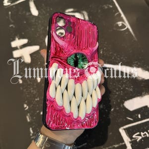 May include: A pink and black phone case with a monster-themed design. The case features a textured, glossy finish with a large set of white teeth, a green eye, and a small white eye. The case has a black border.