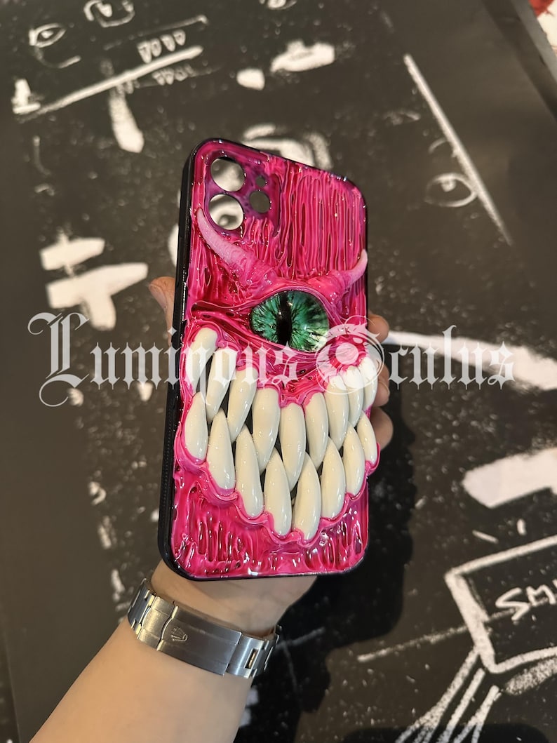 May include: A pink and black phone case with a monster-themed design. The case features a textured, glossy finish with a large set of white teeth, a green eye, and a small white eye. The case has a black border.
