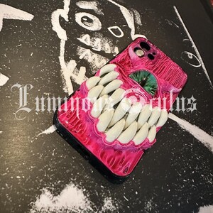 May include: A pink phone case with a grotesque design featuring a mouth with white teeth and a green eye. The case has a glossy finish and is set against a black and white background with the text "Luminous Oculus".