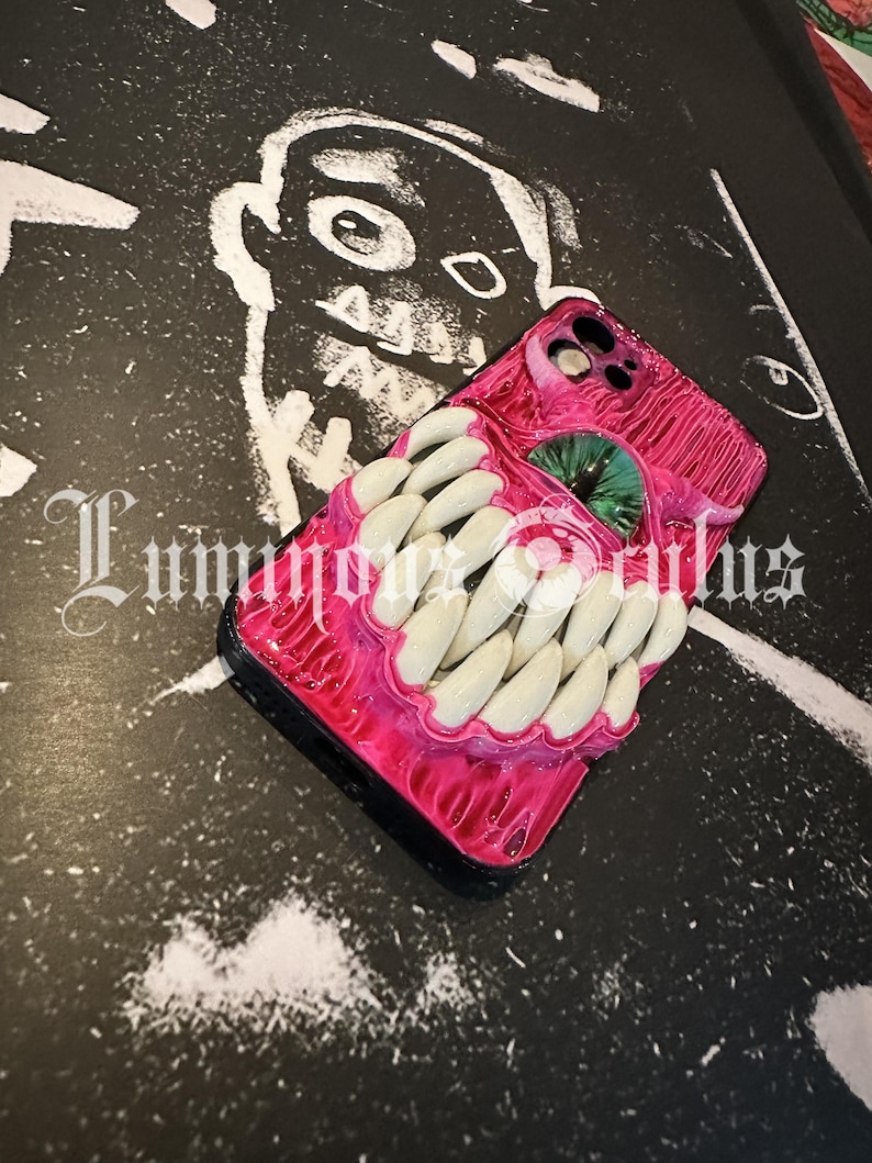 May include: A pink phone case with a grotesque design featuring a mouth with white teeth and a green eye. The case has a glossy finish and is set against a black and white background with the text "Luminous Oculus".