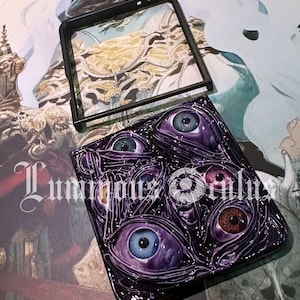 May include: A dark purple phone case with a design of multiple stylized eyes in shades of blue, brown, and purple. The case has a glossy finish and the text "Luminous Souls" is visible. A black frame sits above the case.