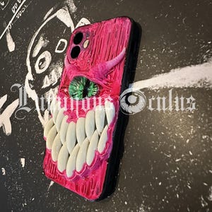 May include: A pink and black phone case with a monster-themed design. The case features a green eye, white teeth, and a horn. The text "Luminous Oculus" is visible on the bottom of the case.