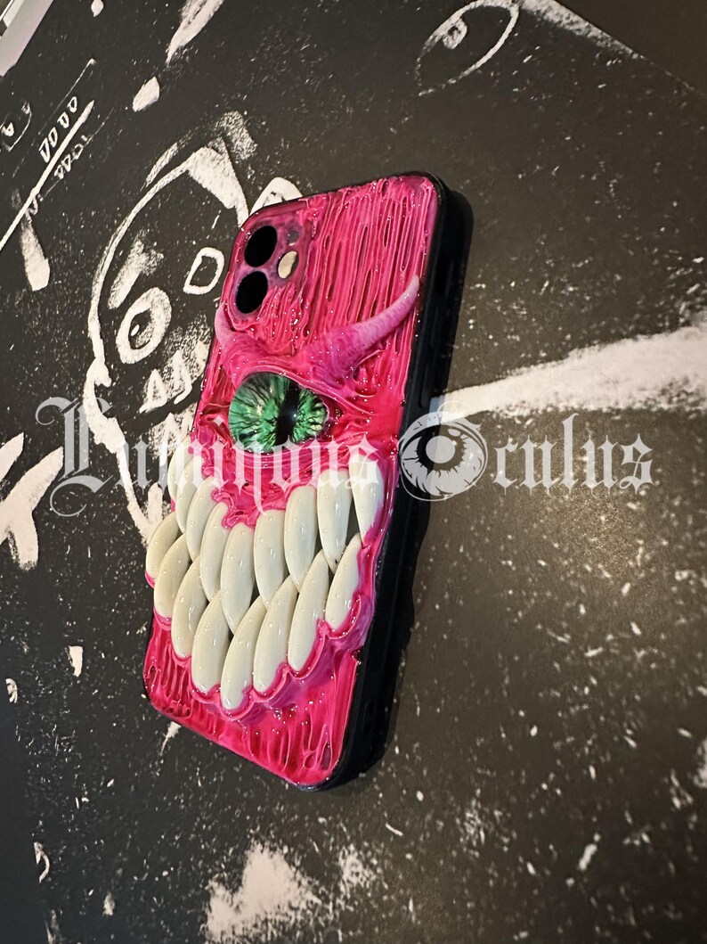 May include: A pink and black phone case with a monster-themed design. The case features a green eye, white teeth, and a horn. The text "Luminous Oculus" is visible on the bottom of the case.