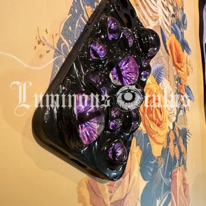 May include: Black phone case with multiple purple and white eye designs. The case has a glossy finish and is mounted on a wall with floral artwork. The text "Luminous Tales" is visible on the lower left.