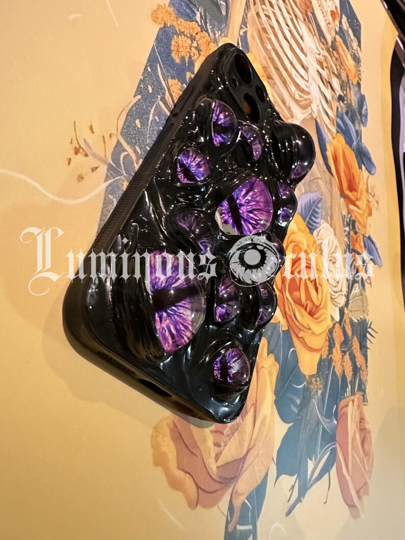 May include: Black phone case with multiple purple and white eye designs. The case has a glossy finish and is mounted on a wall with floral artwork. The text "Luminous Tales" is visible on the lower left.