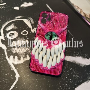 May include: A pink phone case with a monster design. The case features a large eye with a green iris, sharp white teeth, and a textured, glossy finish. The case is on a black surface with white artwork.