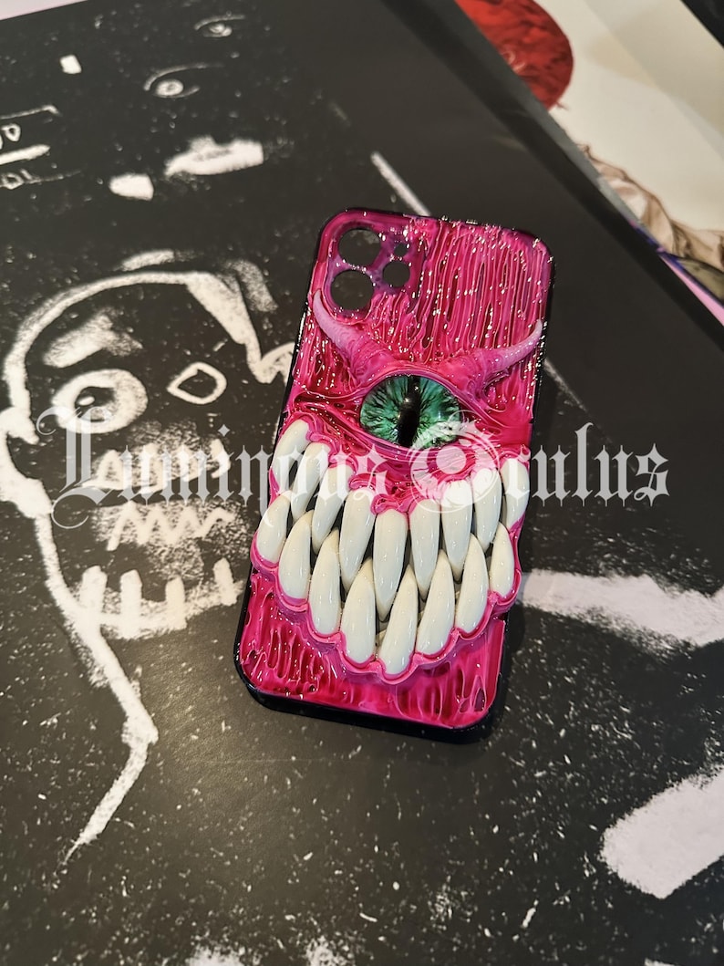 May include: A pink phone case with a monster design. The case features a large eye with a green iris, sharp white teeth, and a textured, glossy finish. The case is on a black surface with white artwork.