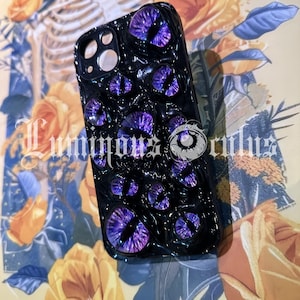 May include: Black phone case featuring multiple purple eye designs. The case has a textured surface and the text "Luminous Orbs". The background includes yellow roses and a skeleton illustration.