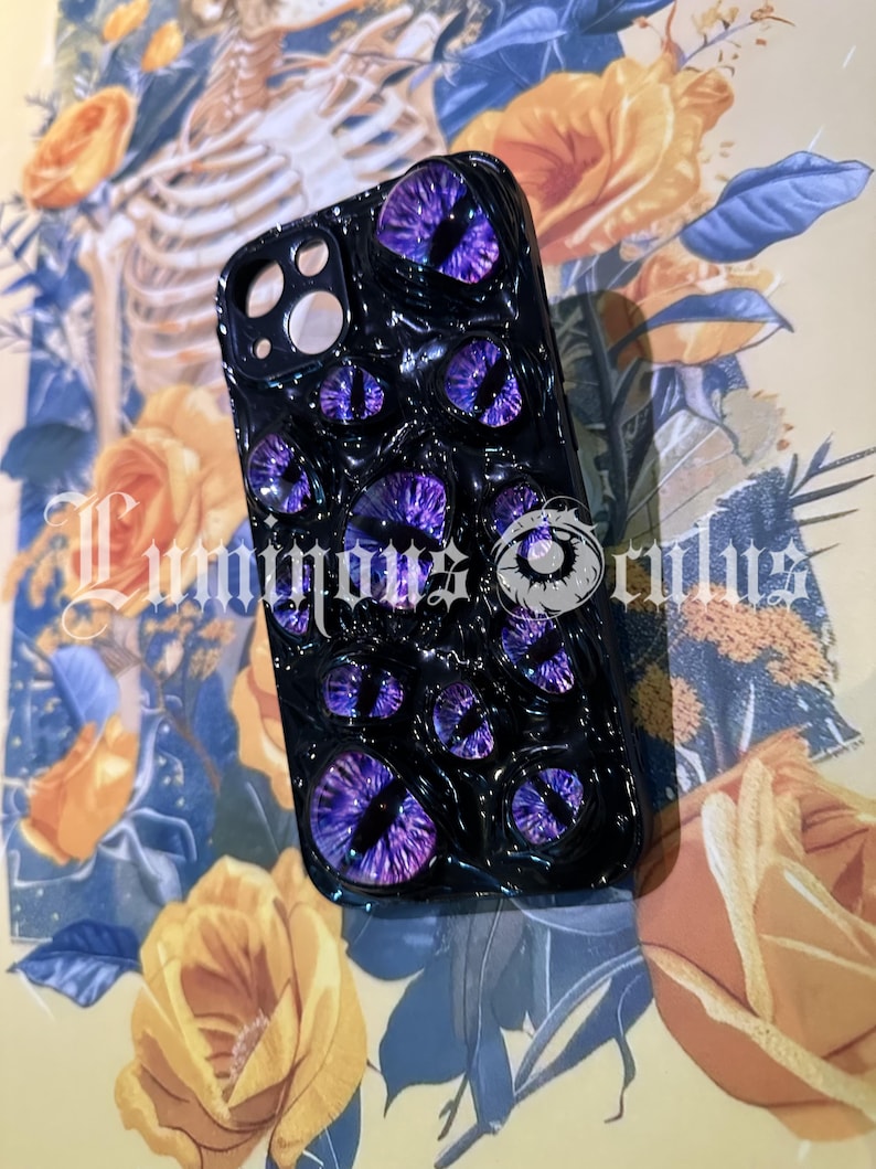 May include: Black phone case featuring multiple purple eye designs. The case has a textured surface and the text "Luminous Orbs". The background includes yellow roses and a skeleton illustration.