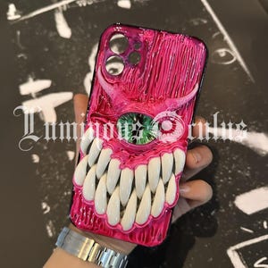 May include: A pink and black phone case featuring a monster design with large white teeth, a green eye, and a textured surface. The case has a glossy finish and a black border.