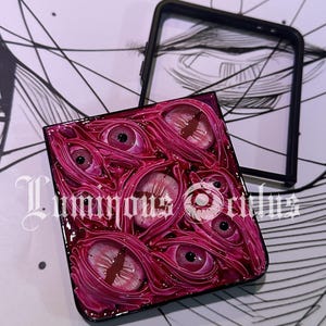 May include: A phone case featuring a detailed, textured design of multiple pink and red eyes. The case has a glossy finish and the text "Luminous Orders" is visible. A black phone case frame is also present.