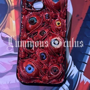 May include: A black phone case with a detailed design of red-veined eyeballs with different iris colours. The case features the text "Luminous Oculus" in a decorative font.