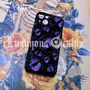 May include: Black phone case featuring multiple purple eye designs. The case has a glossy finish and is set against a backdrop of a skeleton, roses, and floral patterns. The text "Luminous Occults" is visible.