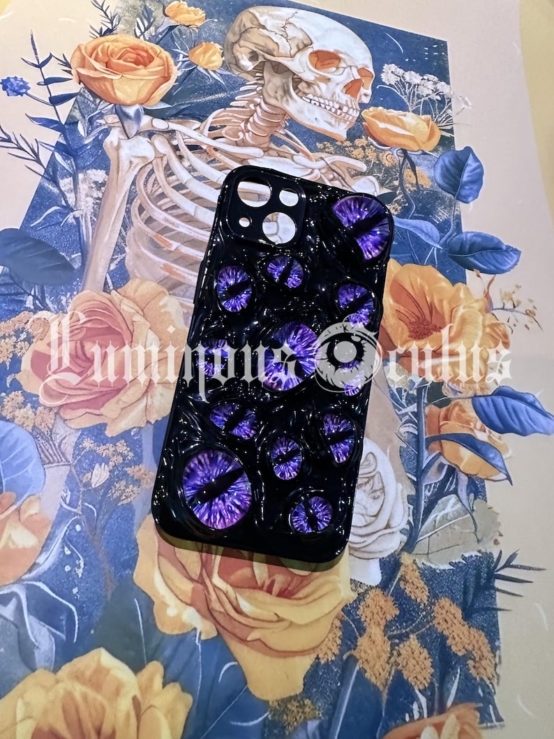 May include: Black phone case featuring multiple purple eye designs. The case has a glossy finish and is set against a backdrop of a skeleton, roses, and floral patterns. The text "Luminous Occults" is visible.