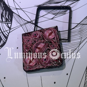 May include: A black phone case with a detailed design of multiple red and pink eyes. The case has the text "Luminous Oculus" in a decorative font. A black frame sits above the case.