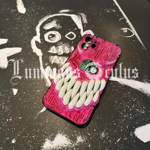 May include: A pink and black phone case with a monster-themed design. The case features a textured, fleshy appearance with a large green eye and rows of white teeth. The background is a black and white illustration.