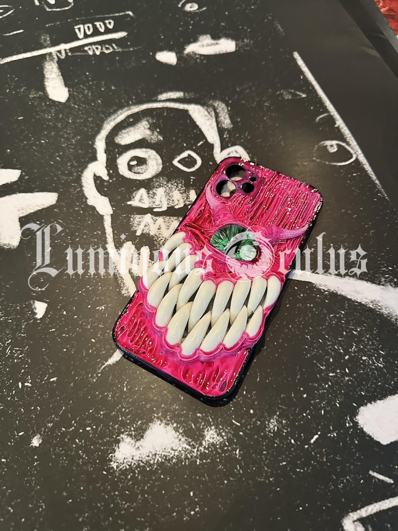 May include: A pink and black phone case with a monster-themed design. The case features a textured, fleshy appearance with a large green eye and rows of white teeth. The background is a black and white illustration.