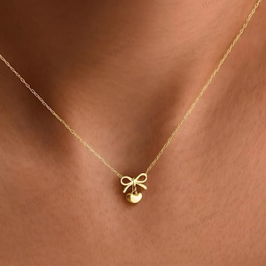 Può includere: Una delicata collana d'oro con un piccolo fiocco e un ciondolo a forma di cuore. La sottile catena sostiene il ciondolo, creando un design semplice ed elegante. La collana è mostrata su uno sfondo color carne.