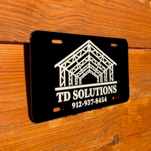 May include: Black license plate with white text and a building graphic. The text reads "TD SOLUTIONS" and a phone number. The plate has three holes for mounting and is displayed against a wood background.