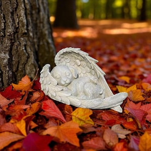 May include: A sleeping puppy sculpture with angel wings, in a serene pose. The statue is light gray and rests among colorful autumn leaves. The sculpture is a decorative item.