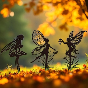 Puede incluir: Tres estacas de jardín de hadas de metal negro. Una hada está sentada en una seta, otra sostiene una cesta y la tercera una linterna. Las estacas están colocadas en un jardín con hojas de otoño.