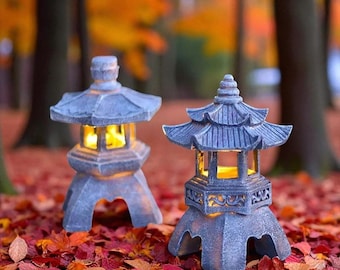 May include: Two gray stone Japanese garden lanterns with pagoda-style roofs. The lanterns are lit from within, casting a warm glow. They sit on a bed of red and orange autumn leaves, with a blurred forest background.